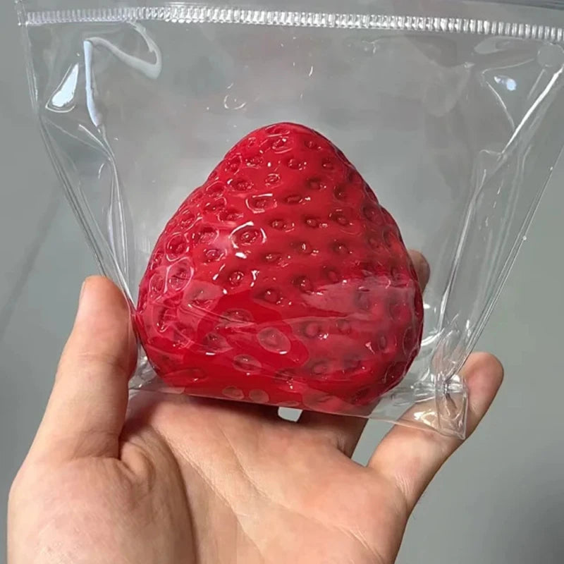 A hand holds a bright red strawberry squeeze fidget toy sealed in a clear plastic bag.