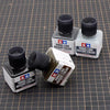 Three glass bottles of Panel Line Accent Color enamel paint in black and gray shades resting on a grid surface.