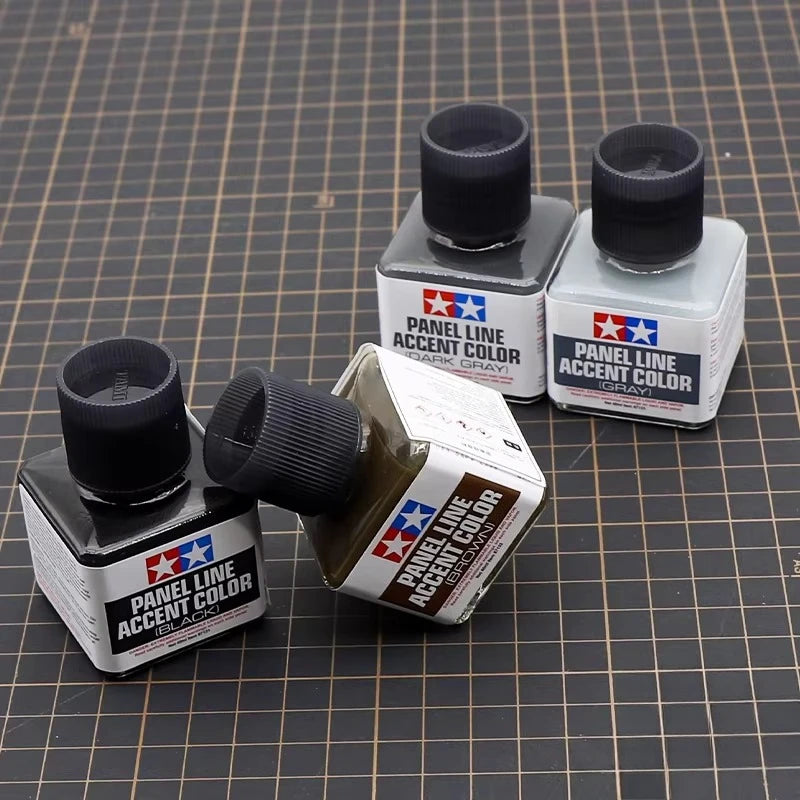 Three glass bottles of Panel Line Accent Color enamel paint in black and gray shades resting on a grid surface.