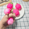 A hand is holding and squeezing a bright pink dragon fruit squishy stress relief toy on a white plate.