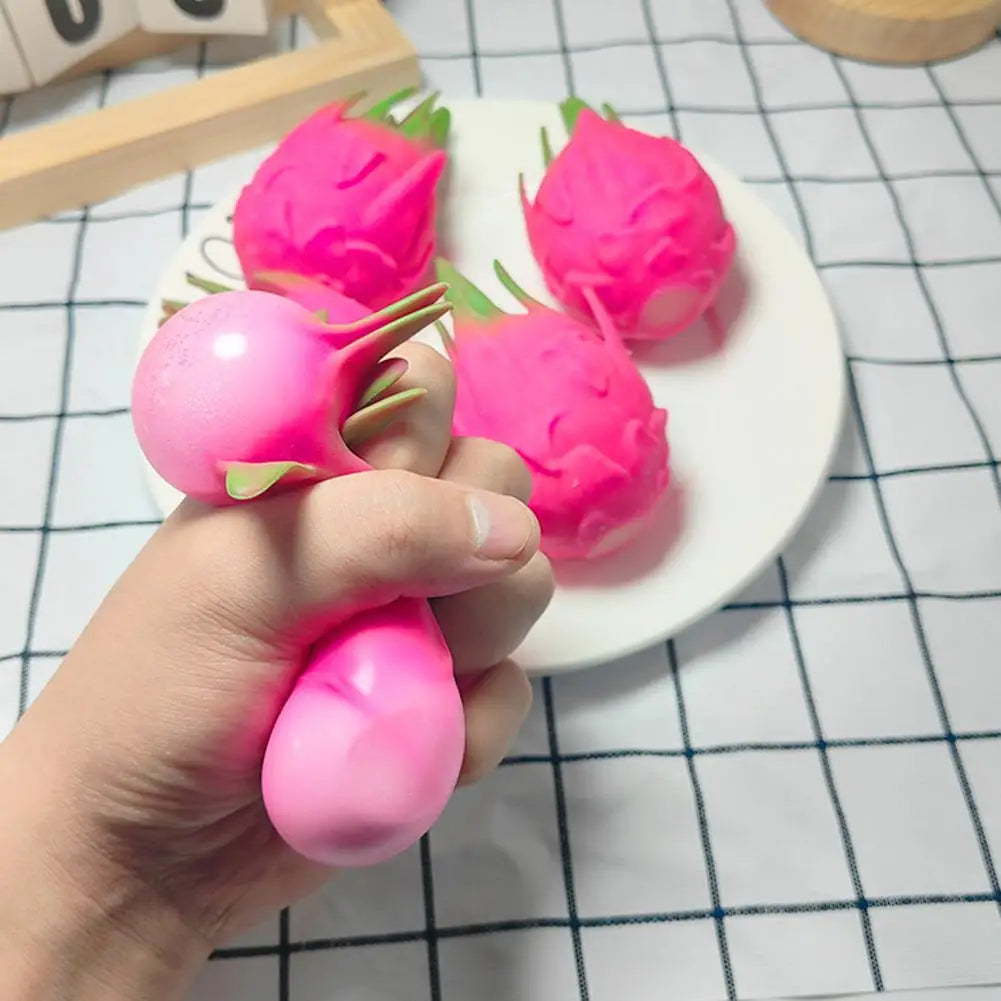 A hand is holding and squeezing a bright pink dragon fruit squishy stress relief toy on a white plate.