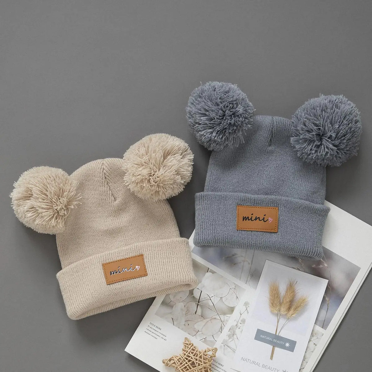 A beige knit baby beanie hat with a fluffy pom-pom and a gray version displayed on a white background.