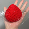 A bright red strawberry squeeze fidget toy is held in a hand, packaged in clear plastic.