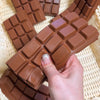 A hand holds a realistic brown chocolate squishy toy with a waffle pattern against a woven background.
