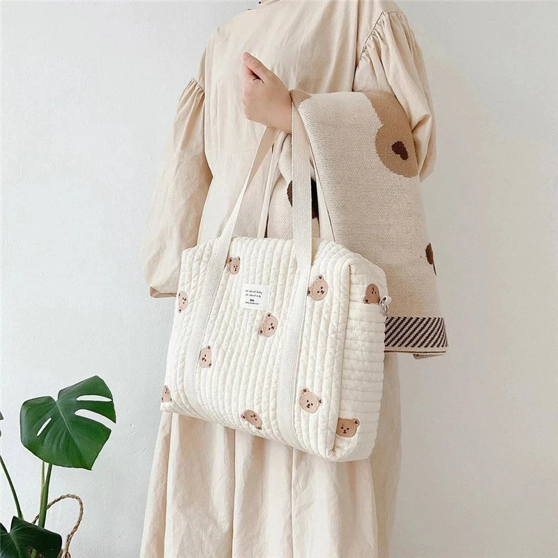 A beige maternity dress is shown holding a white quilted diaper bag with brown embroidery.