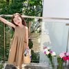 A young girl wearing a beige sleeveless cotton sundress stands outdoors next to a glass railing.