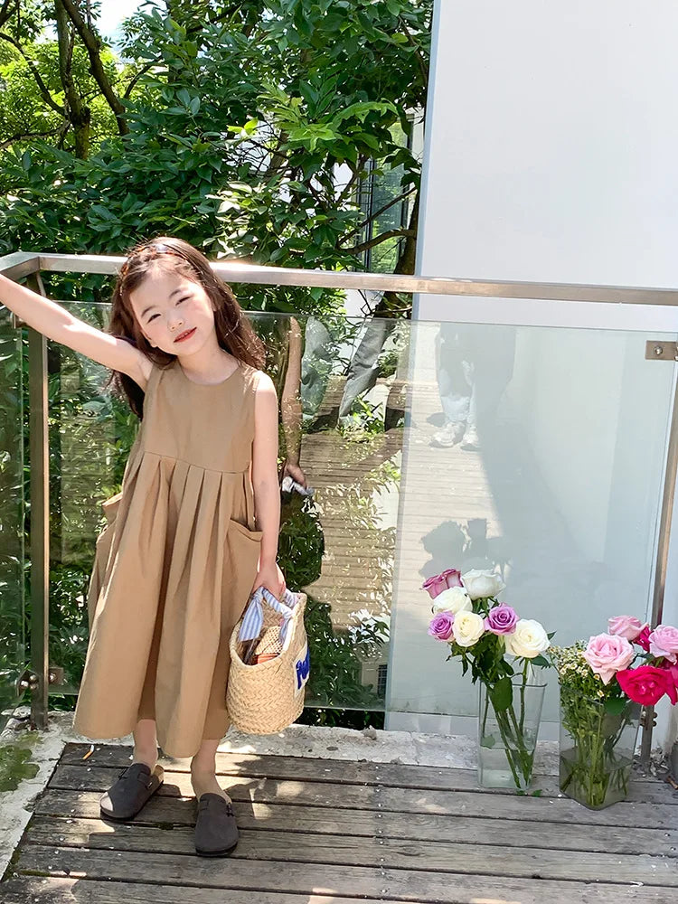 A young girl wearing a beige sleeveless cotton sundress stands outdoors next to a glass railing.