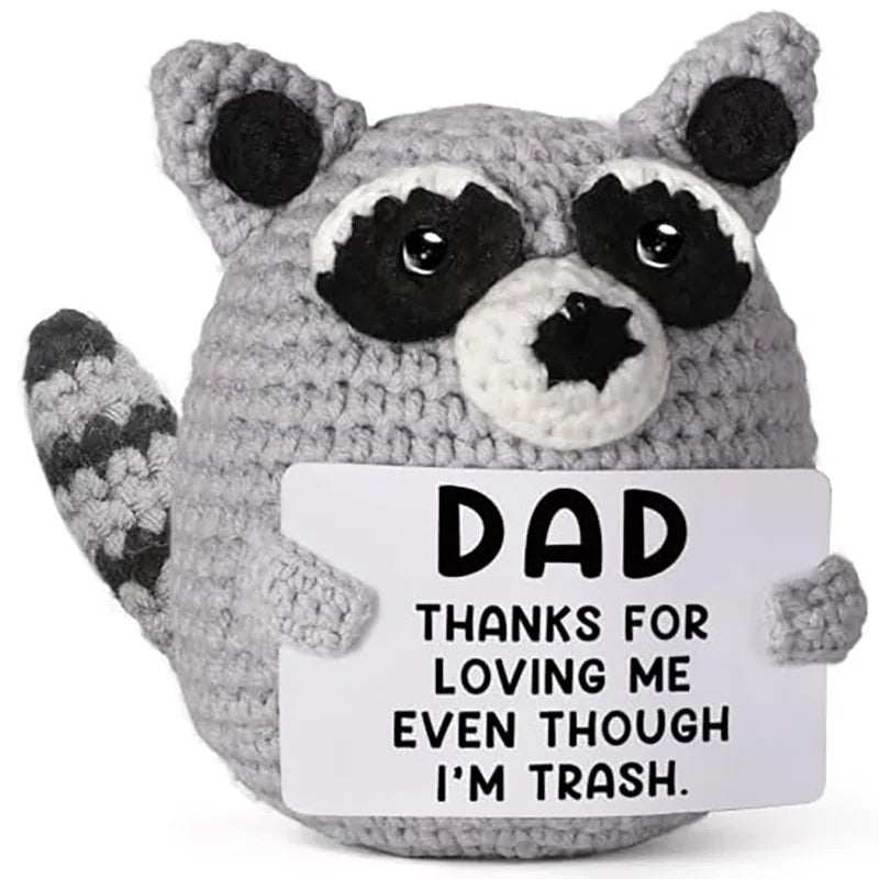 A gray crochet plush toy shaped like a raccoon with black markings, featuring a tag that reads Dad thanks for loving me even 