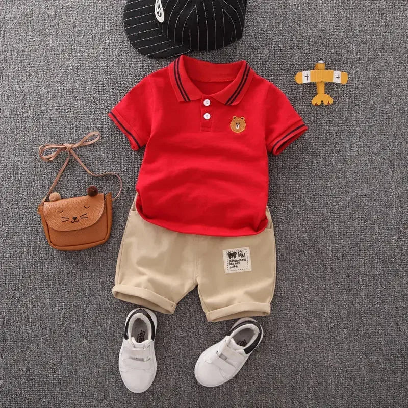 A red polo shirt and beige shorts set for a boy, with a small wooden animal charm nearby.