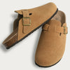 A pair of tan suede cork mules with leather straps and arch support shown from the side.