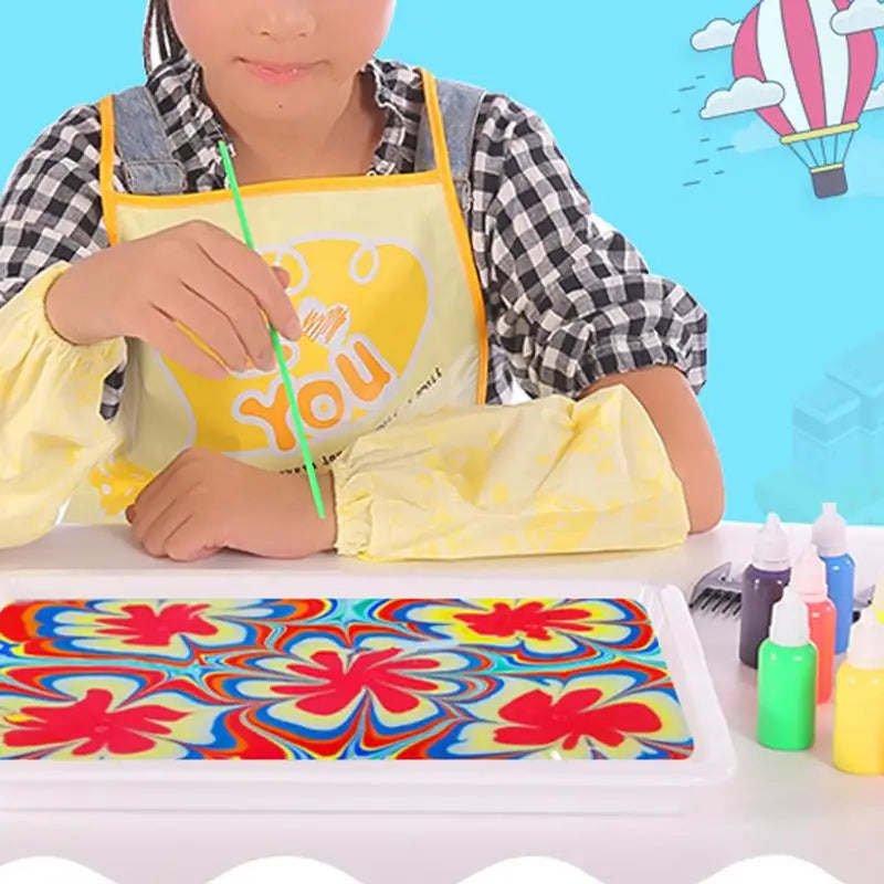 A child wearing an apron is painting a colorful floral design on a white tray with water marbling paint.