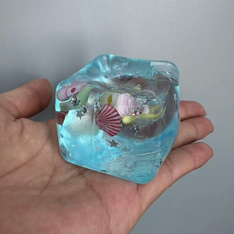 A hand holds a translucent blue ice-like block with various colored inclusions and shell shapes.