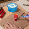 A hand is using a blue wax seal melting kit with a heater and spoon to melt red wax on brown paper.