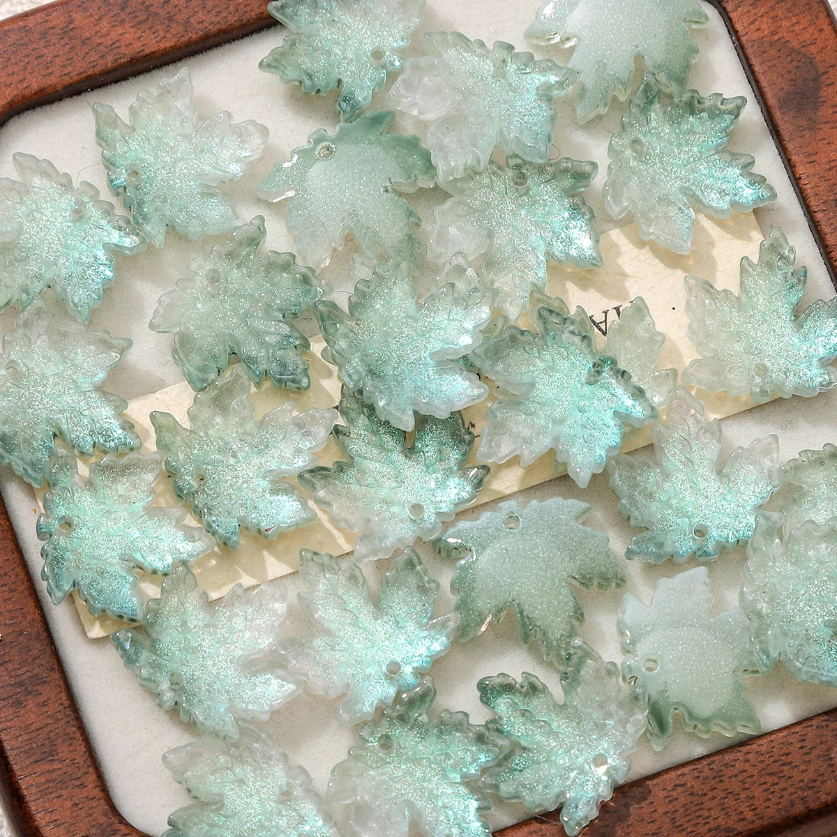 Clear green maple leaf glass beads arranged in a wooden box for DIY jewelry making