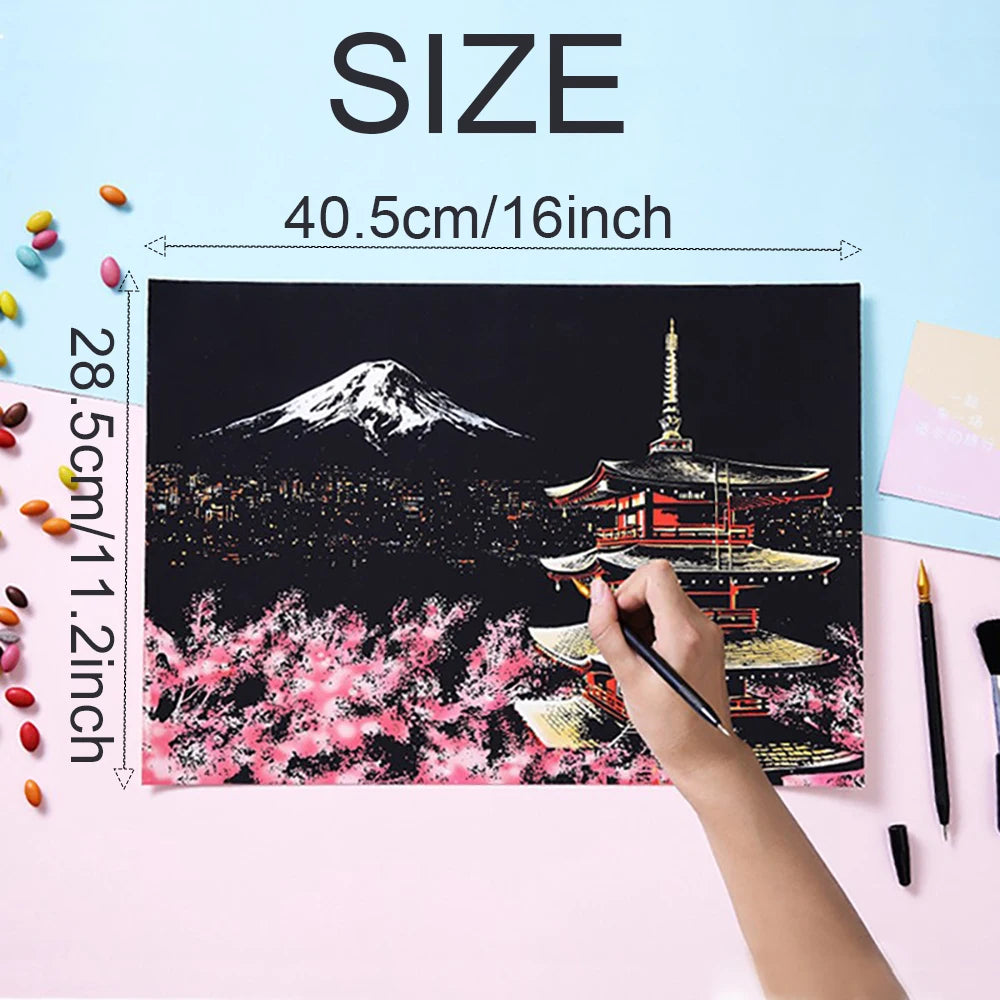 A black scratch art painting depicting Mount Fuji and a cityscape at night, being painted by a hand.