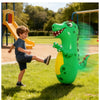 A happy boy is playing with a large green inflatable dinosaur punching bag outdoors on a sunny day.