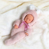 A realistic soft vinyl baby doll wearing a pink fuzzy outfit and hood, lying on white fur fabric.