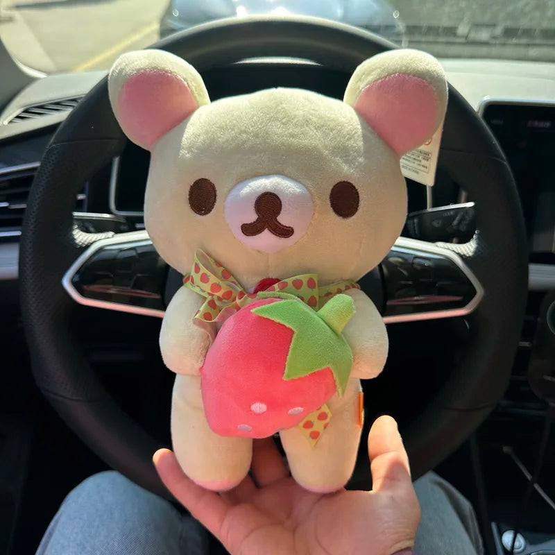 A hand holds a plush toy of Rilakkuma in the form of a strawberry bear with pink and green accents.