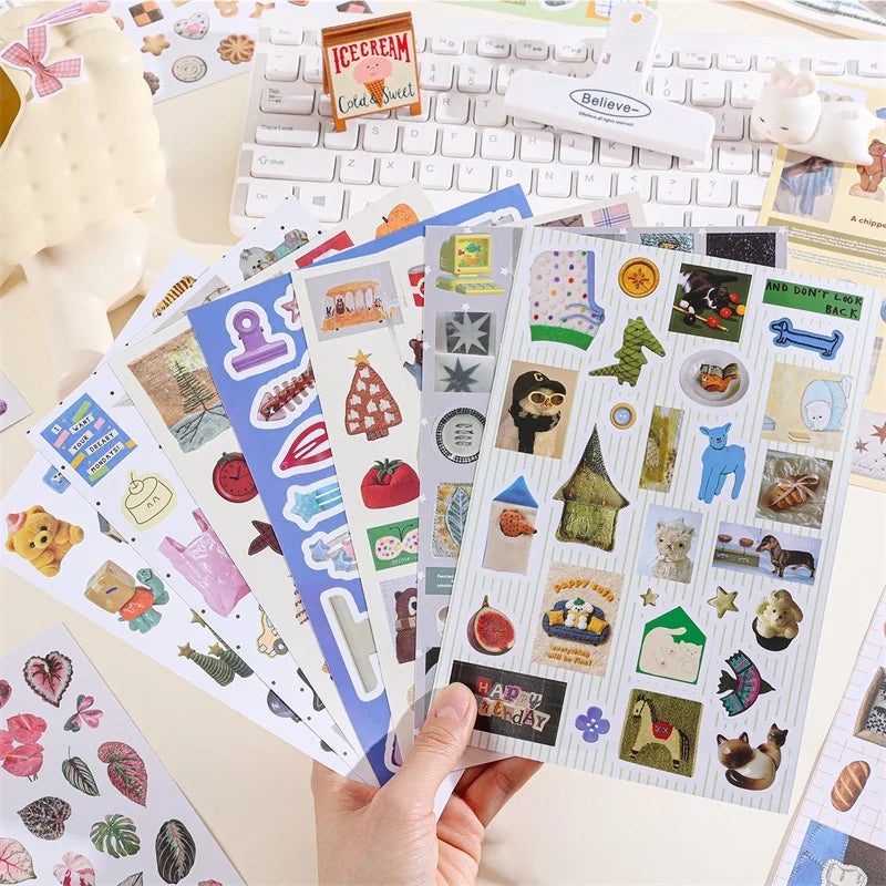 A hand holds an open book of retro aesthetic stickers for DIY crafting on a white desk.