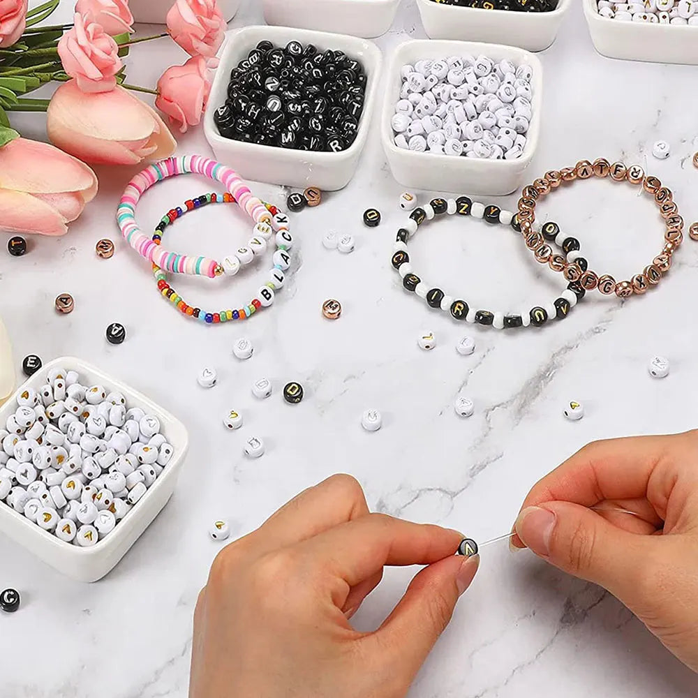 A hand is assembling jewelry using small acrylic alphabet and number beads scattered on a white marble surface.