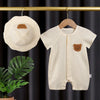 A beige cotton muslin baby romper and hat set displayed hanging against a dark curtain.