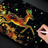 A hand draws a glowing deer in scratch art on a dark surface with scattered gold and green glitter.