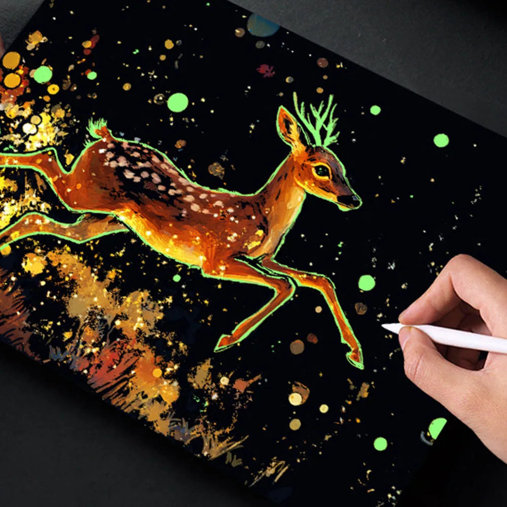 A hand draws a glowing deer in scratch art on a dark surface with scattered gold and green glitter.