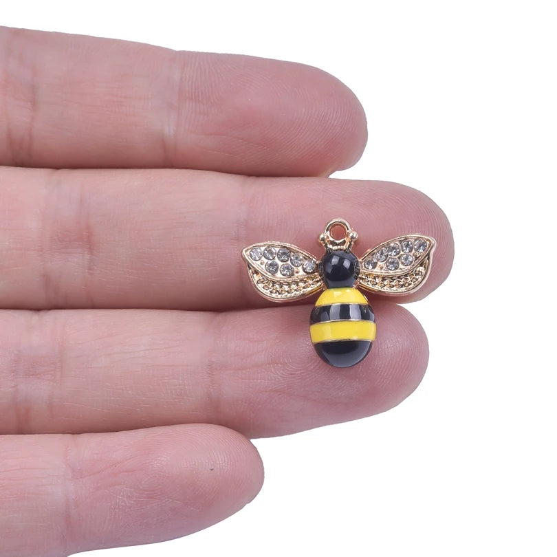 A pair of gold-toned bee enamel charms with black and yellow stripes and sparkling wings are held on a person's finger.