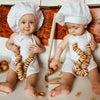 Two babies wearing white chef hats and aprons sitting on a surface with wooden cooking tools.