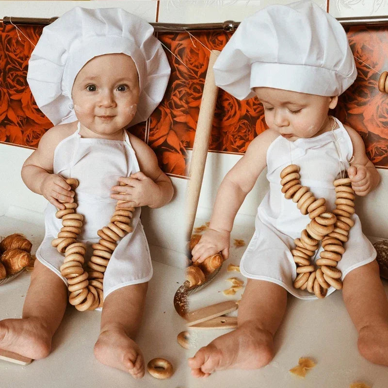 Two babies wearing white chef hats and aprons sitting on a surface with wooden cooking tools.