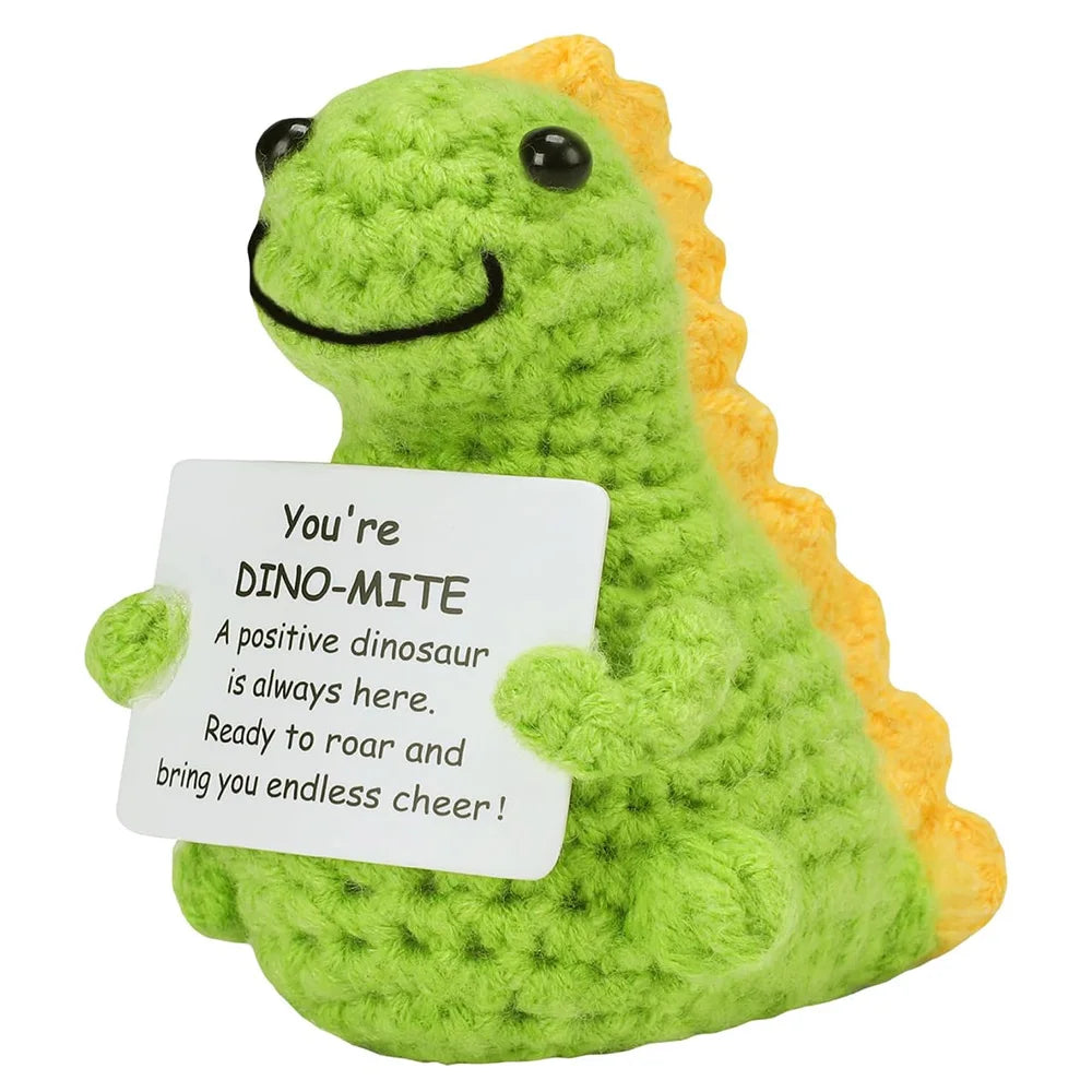 A handmade crochet dinosaur toy with a smiling face and yellow spines, holding a sign that reads Dino-mite.