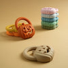 A set of silicone pumpkin baby teethers in orange and light green colors are displayed on a beige surface.