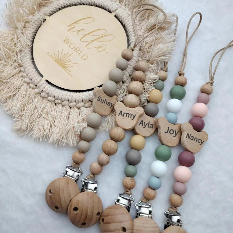 A set of personalized wooden baby pacifier clips with silicone beads hanging from a natural fiber wreath.