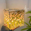 A clear cube night light filled with glowing yellow and orange tulip-shaped lights, held in a hand against a white background