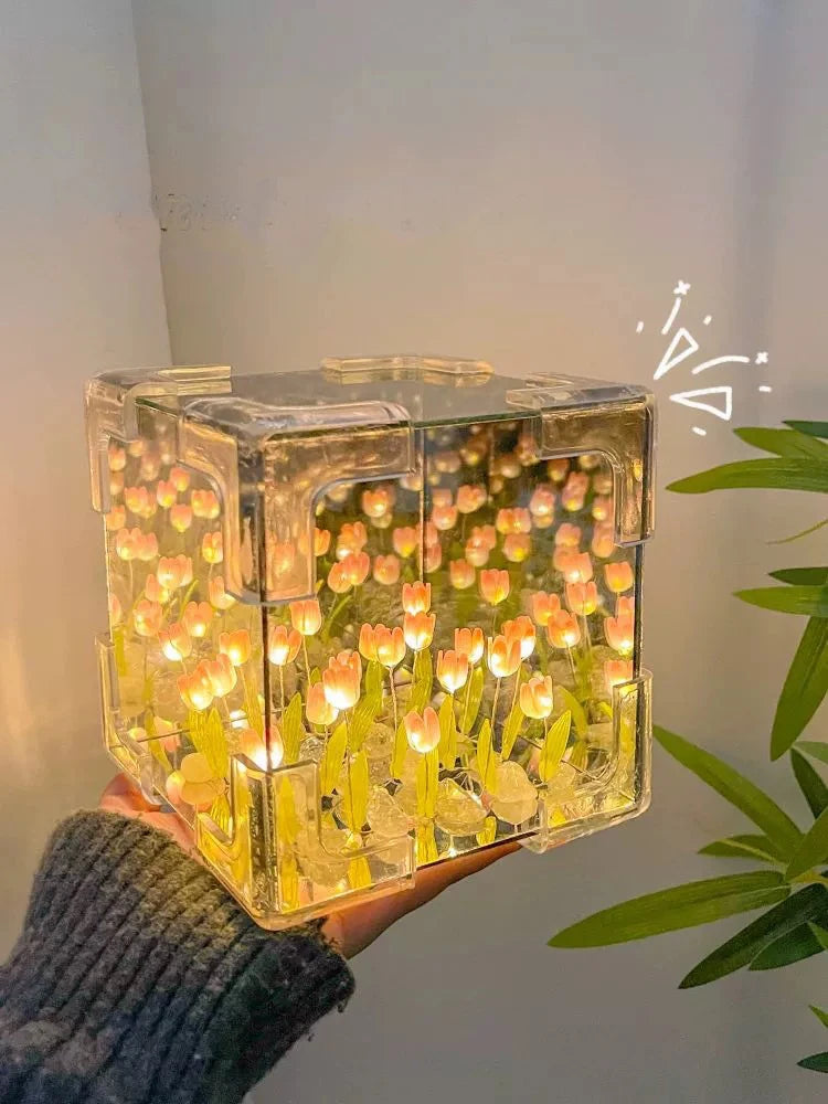 A clear cube night light filled with glowing yellow and orange tulip-shaped lights, held in a hand against a white background