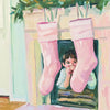 A pair of pink boots are displayed hanging on a wooden shelf, with green foliage in the background.
