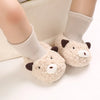 A pair of soft, light beige cartoon baby booties with brown accents and knitted socks shown on a person's foot.