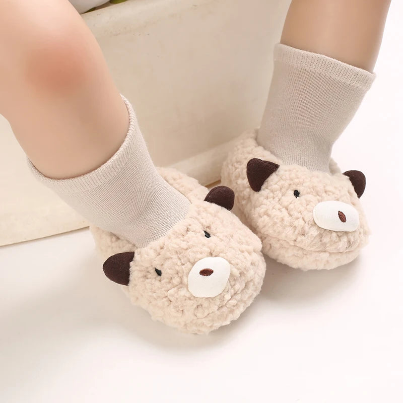 A pair of soft, light beige cartoon baby booties with brown accents and knitted socks shown on a person's foot.