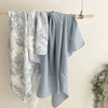 A light blue muslin swaddle blanket with a floral print hanging on a wooden rack.