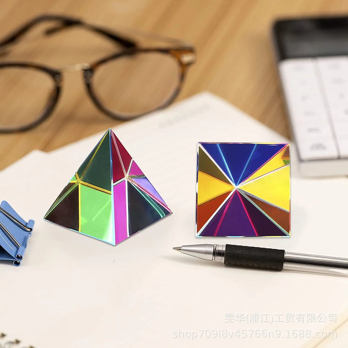 A geometric prism cube with iridescent colors rests on a white surface next to a pen and eyeglasses.