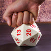 A hand holds a white D20 polyhedral dice on a dark surface.