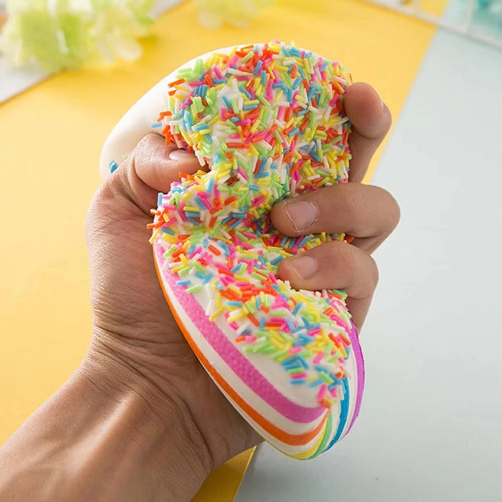 A hand holds a colorful rainbow layer cake squishy toy with sprinkles against a white background.