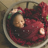 A newborn baby is swaddled in a deep red knit wrap with a pearl headband, resting in a woven bowl.