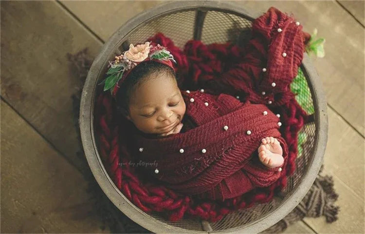 A newborn baby is swaddled in a deep red knit wrap with a pearl headband, resting in a woven bowl.
