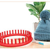 A teal knitted hat and a red round knitting loom with crochet hooks are displayed on a wooden surface.