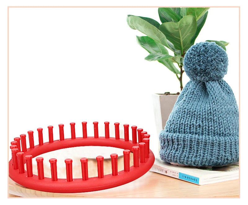 A teal knitted hat and a red round knitting loom with crochet hooks are displayed on a wooden surface.
