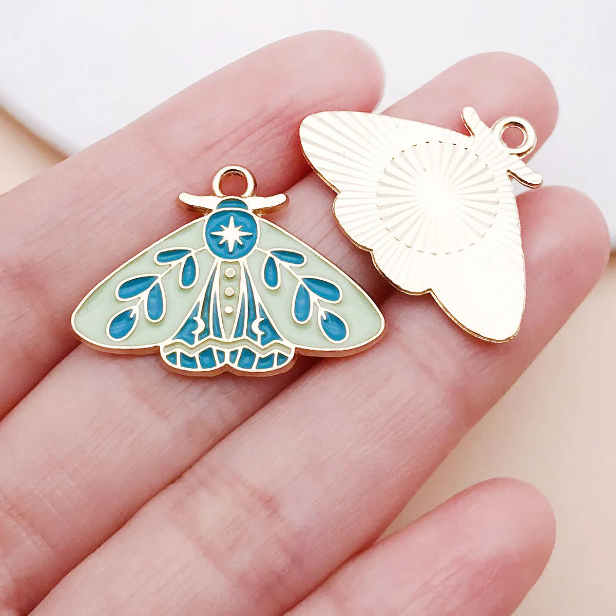 Gold-toned butterfly drop charm with iridescent blue and green enamel details shown on a hand.