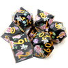 A set of seven black resin polyhedral dice with gold numbers and floral designs displayed on a white background.