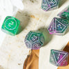 A collection of seven iridescent resin polyhedral dice set on a light marble surface.