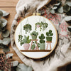 A DIY cross stitch starter kit featuring green potted plants embroidered on a circular wooden board.